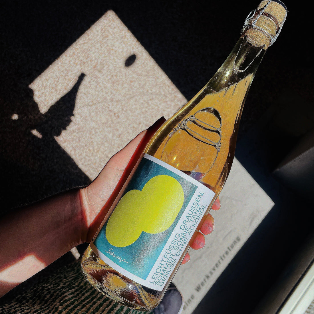 In der linken Hand eine geschlossene 0,75 ltr. Flasche Sparkling Tea mit dem Namen DITO Soberitif von Weingut Kern aus Württemberg.